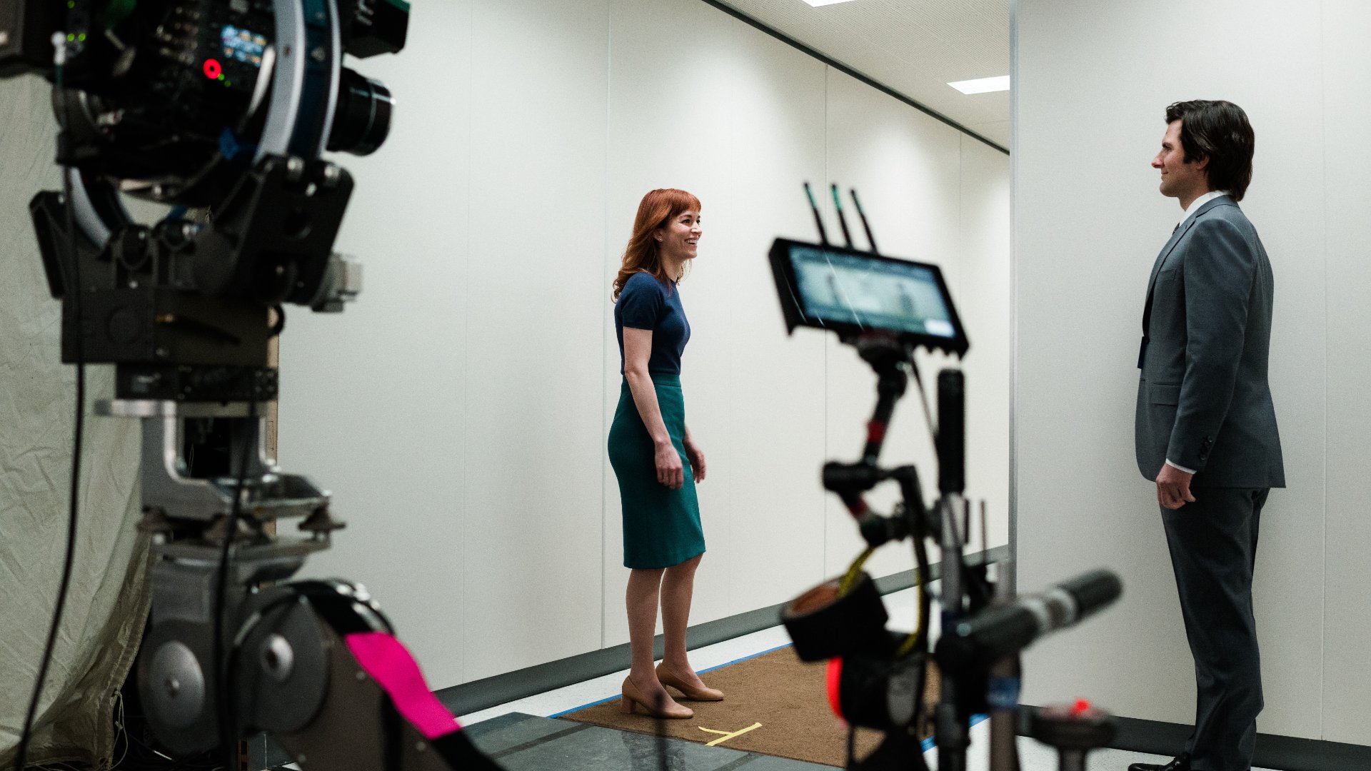 BTS image of Britt Lower as Helly and Adam Scott as Mark on the set of Severance. They are on a film set featuring a professional camera setup focusing on them inside a minimalist, modern office environment. Britt Lower is dressed in a form-fitting knee-length dress, smiles as she stands facing Adam Scott, who is clad in a formal suit and appears relaxed. The scene is monitored by a digital display screen connected to the camera, showing live feedback of the shot being captured. The overall setup suggests a dialogue or interaction scene being filmed, highlighting the actors' expressions and positioning in a controlled, clean setting.