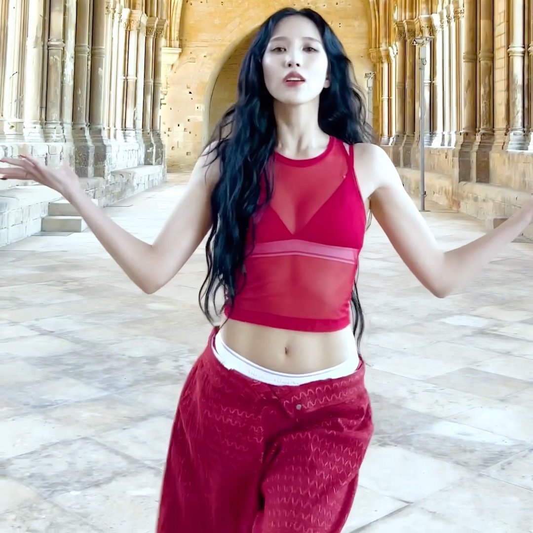 A woman with long dark hair wearing a red mesh crop top and red velvet pants, posing with arms outstretched in a gothic architectural corridor.