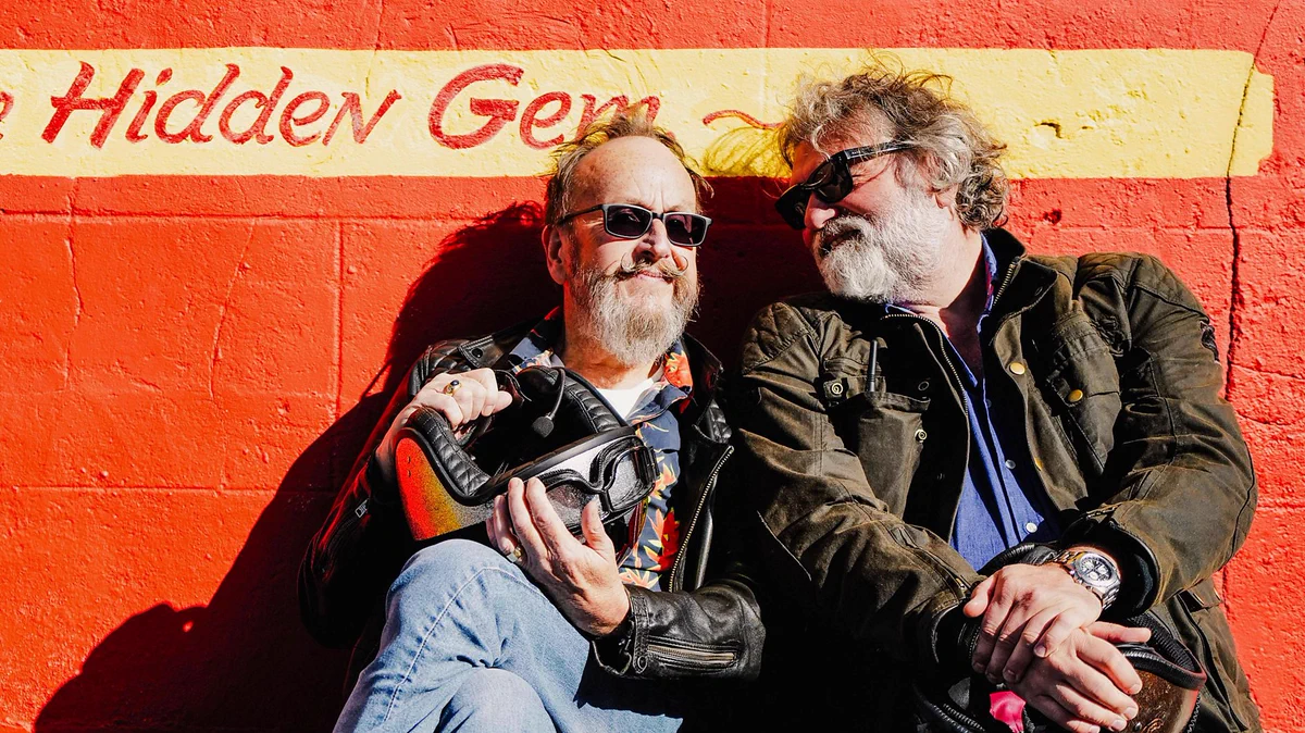 Dave Myers and Si King sitting in their biking gear, leaning against an orange wall and smiling in the sun