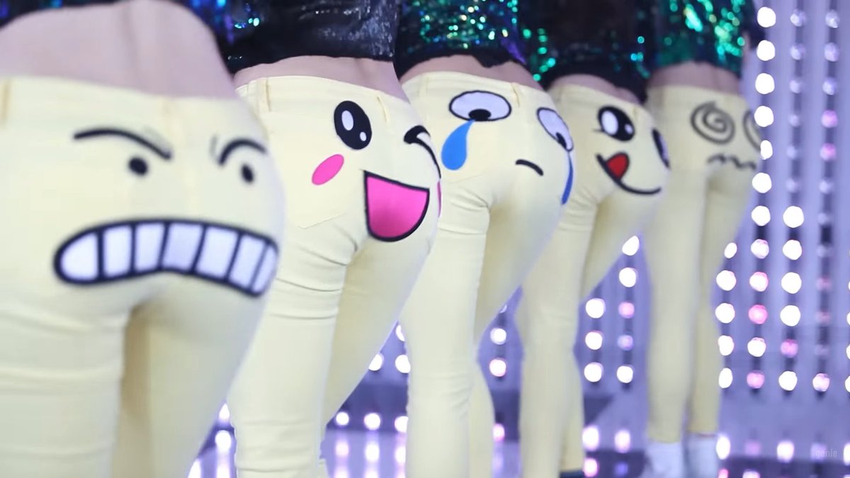 A row of dancers wearing cream-colored leggings with various cartoon emoji-style faces printed on the back (showing expressions like gritted teeth, happy, crying, dizzy, and surprised), performing on a stage with bright LED lights.