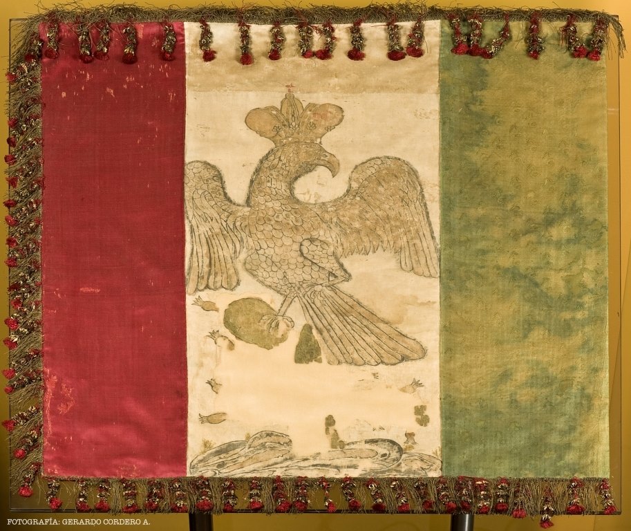 #undíacomohoy en 1821 se decretó que la bandera del Imperio Méxicano sería tricolor: verde,blanco y rojo,con franjas verticales y al centro la efigie de un águila coronada, sin serpiente, posando sobre un nopal nacido en un islote, retomando el símbolo fundacional de Tenochtitlan