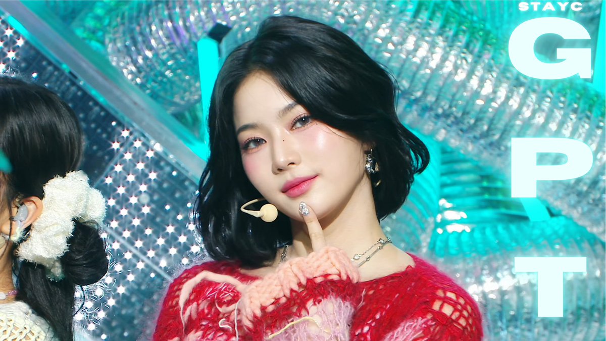 A stage performance image of a K-pop idol with shoulder-length dark hair, wearing a red fuzzy outfit and a headset microphone, with "STAYC" and large letters "G", "P", "T" on the right side.