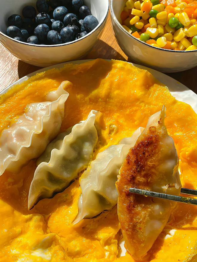 A delightful fusion of crispy dumplings nestled on a cloud of fluffy scrambled eggs, with fresh blueberries and a vibrant veggie medley adding bursts of color and flavor. A perfect way to brighten up your meal!