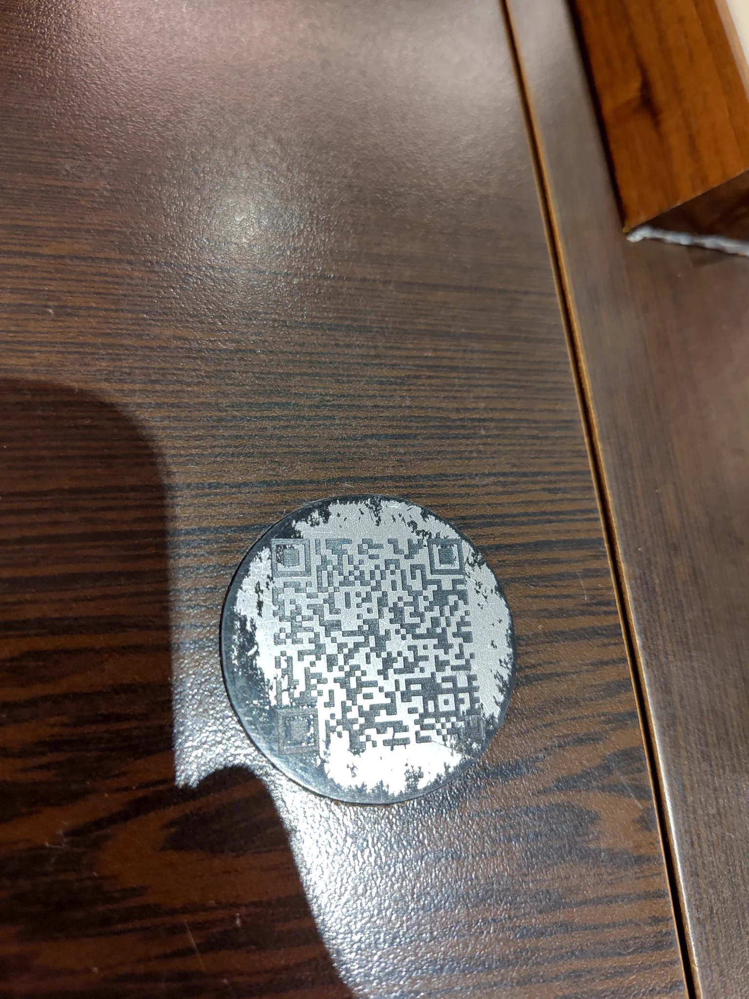 A QR code affixed to a restaurant tabletop as a black plastic disc with silver paint. The silver paint is badly worn.