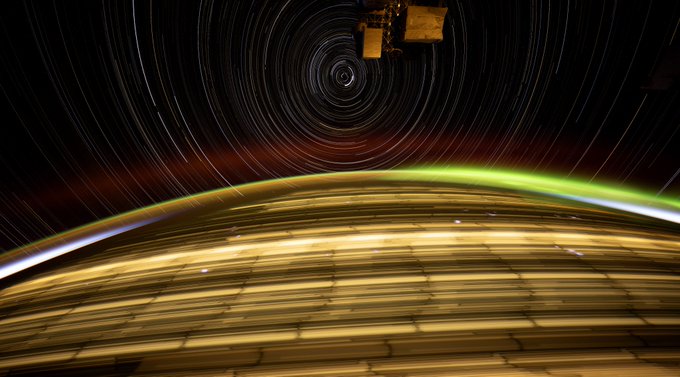 View of Earth from space. The curve of Earth fills the bottom half of the frame, streaks of horizontal golden lights covering its otherwise dark surface. Earth’s atmosphere glows with light blue slivers on either side of the frame, with hues of green and orange in the middle. Above the Earth, there are streaks of light in a radial pattern, glowing brilliantly against the darkness of space. A piece of the ISS is seen at the top of the frame.