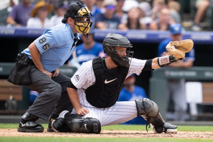 Jacob Stallings reaches out with his glove to catch a ball from his stance behind home plate. He is wearing his catcher gear and the white pinstripe Rockies uniform. Directly behind him is the umpire wearing a blue shirt, grey pants and an umpire mask.