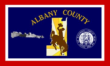 The Albany County, Wyoming flag features a blue field with a thick white and red border, mirroring the state flag. It displays the county's outline, a rider on horseback, the county's name at the top, the county seal on the right, and the Laramie Mountains on the left.