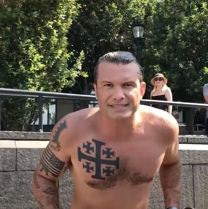 The image shows Pete Hegseth, Donald Trump\'s nominee for Defense Secretary, shirtless and displaying tattoos on his chest and arms. A prominent Jerusalem cross is visible on his chest, and his arm features tattoos including an American flag, Hebrew text, and the Latin phrase "Deus Vult" in Gothic script, alongside a sword and serpent. The setting appears to be an outdoor urban area with greenery, a stone wall, and people in the background. The post text explains the tattoos\' religious and political significance, linking them to evangelical Zionism, end-times crusades, and potential military actions, referencing a NYPost article.