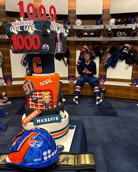The hard hat and championship belt are on a table in front of a cake while Connor McDavid sits in his stall in the Oilers locker room 