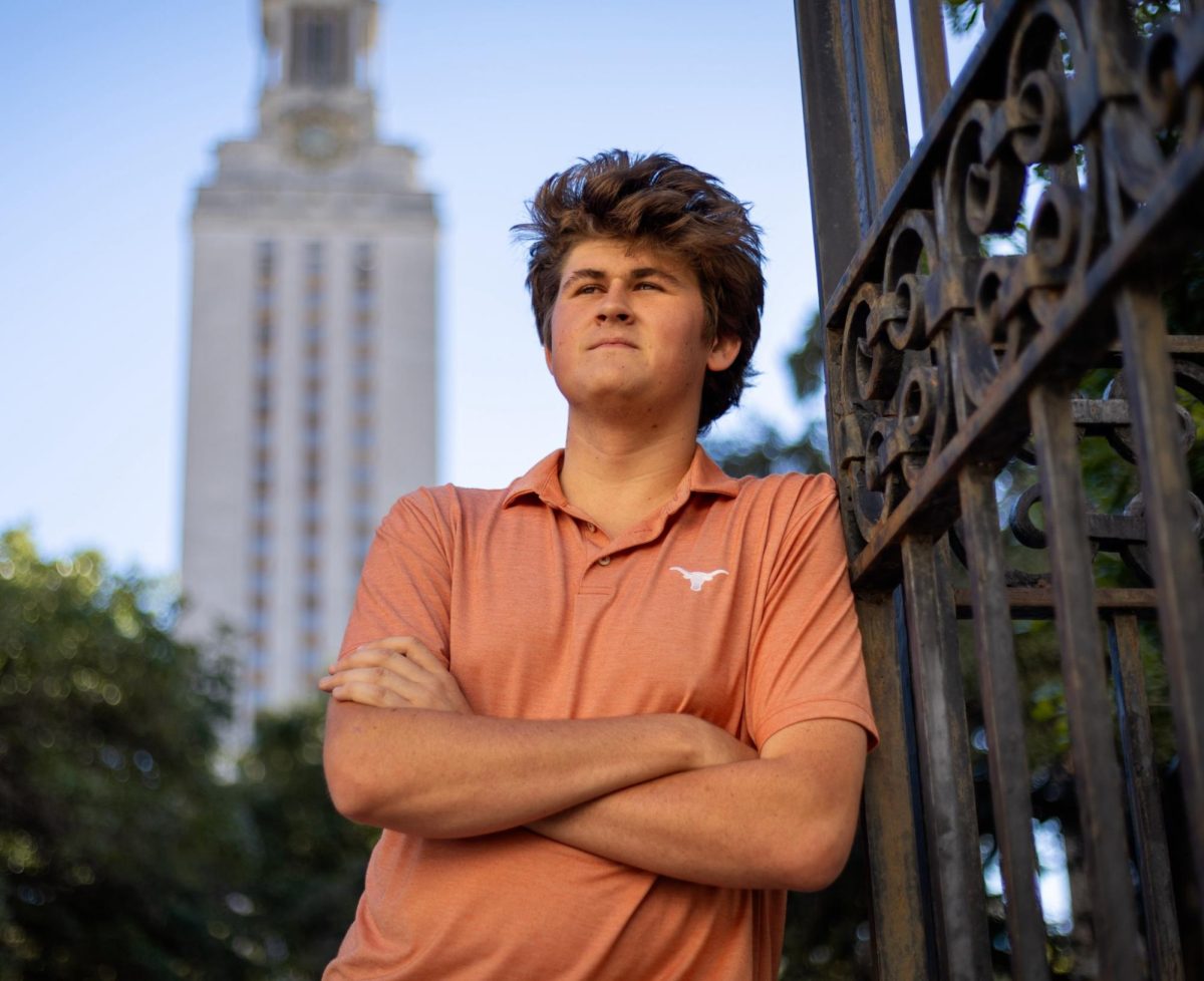 Photo of Grant Walther leaned against a metal gate wearing a burnt orange polo with a longhorn symbol on it. The UT tower stands in the background. Photo by Kamryn Morales.