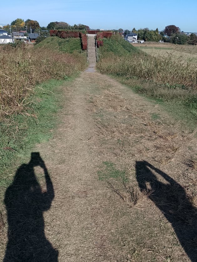 さきたま古墳群の古墳とその中央に設置された階段。両側には草が生い茂る。手前には青空の下で撮影者と同行者の影が映る。