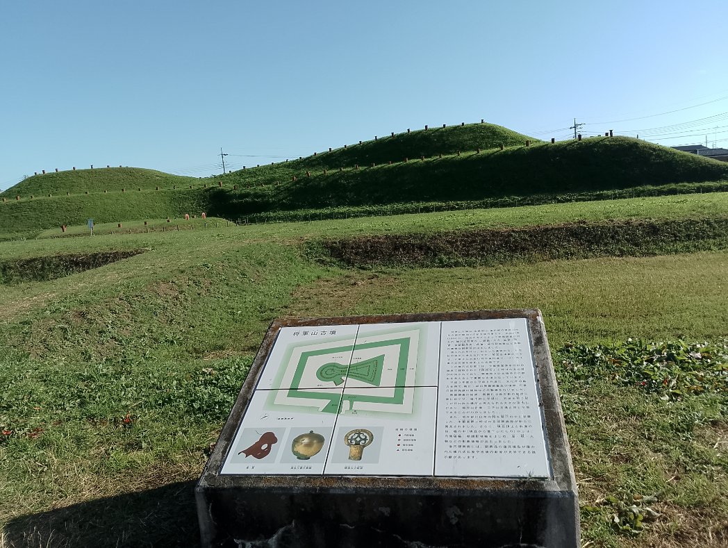 さきたま古墳群の前方後円墳と案内板。案内板には古墳の形状図と埴輪、勾玉などの出土品の説明が記載されている。青空の下、芝生の緑が広がる風景。