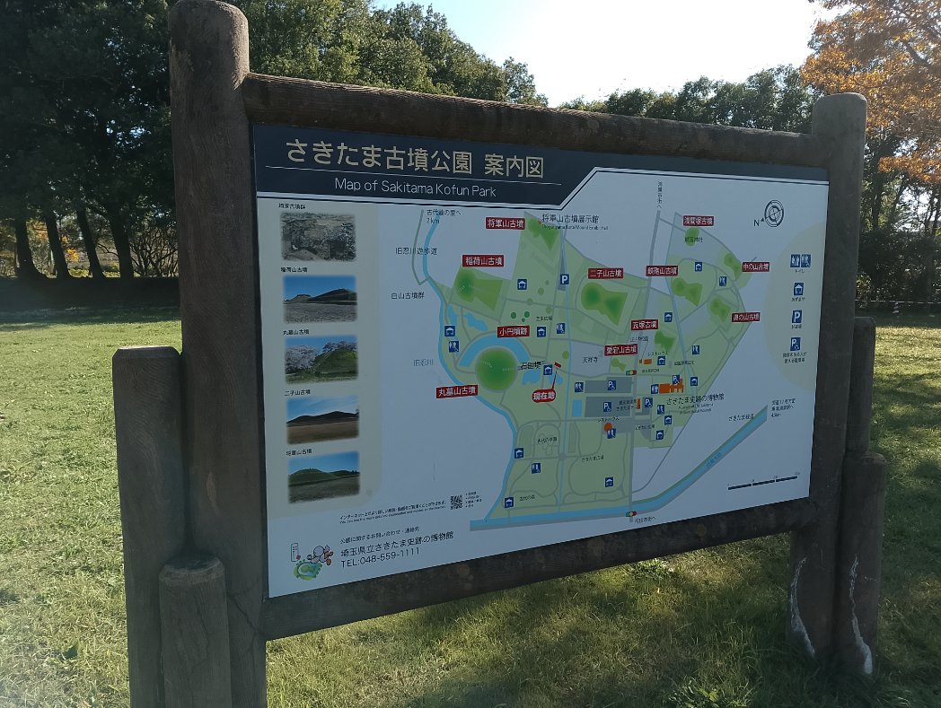 さきたま古墳公園の案内図の看板。公園内の古墳の位置や駐車場、観光スポットが示されている。背景には木々と青空が広がる。