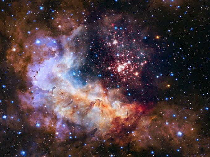 A vibrant image of the Westerlund 2 star cluster surrounded by the colorful, swirling cloud forms of the Gum 29 nebula. Bright stars illuminate the reddish and blue nebulous regions.