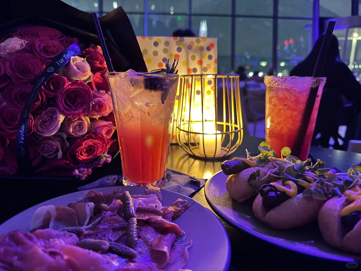 orientalli's tweet image. A romantic dinner setting with a bouquet of roses, refreshing drinks, and delicious appetizers under ambient lighting.