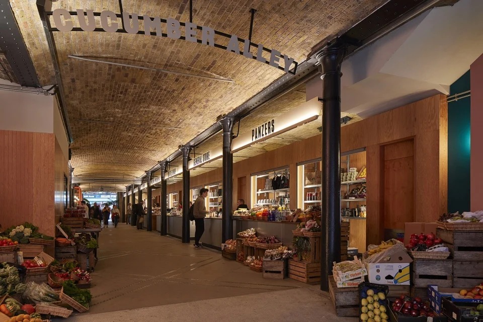 orientalli's tweet image. A bustling market area under an arched ceiling with stalls selling fresh produce and goods, adorned by a sign reading "Cucumber Alley