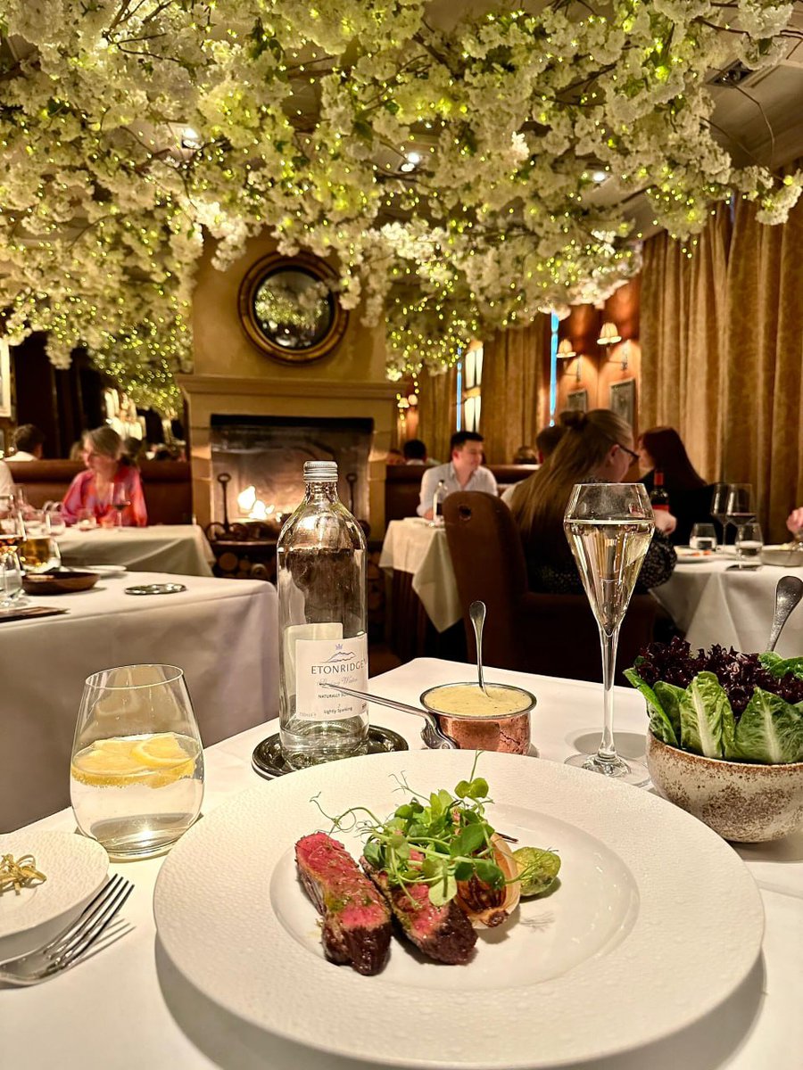 orientalli's tweet image. A beautifully designed menu for Clos Maggiore held under a ceiling adorned with cascading floral decorations and twinkling lights.