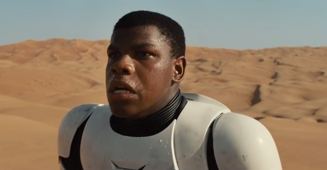 A still from the first trailer for The Force Awakens, showing Finn, a black man with short hair wearing white armour in the hot Jakku sun. Behind him are sand dunes far into the distance and a muddy blue sky. Beads of sweat are forming on his face as he looks flustered.