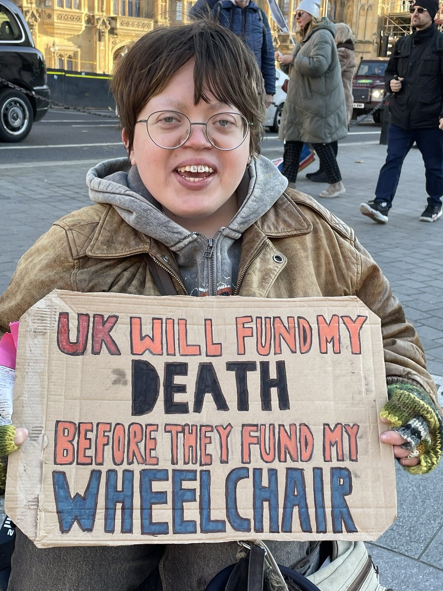 #AssistUsToLive 

Rob holds her placard sitting in her wheelchair which says UK will fund my death before they fund my wheelchair