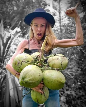 The image features a woman, presumably a vegan influencer, holding a large bunch of green coconuts in one hand while flexing her other arm to show her muscles. She is wearing a black hat, a black tank top, and jeans, standing in what appears to be a natural, outdoor setting with greenery in the background. The post text reveals that this influencer, known for promoting a fruit-only diet, tragically passed away at 39 due to malnutrition after following this diet for 10 years. The image contrasts her apparent physical strength with the underlying health issues caused by her dietary choices, highlighting the dangers of extreme diets.