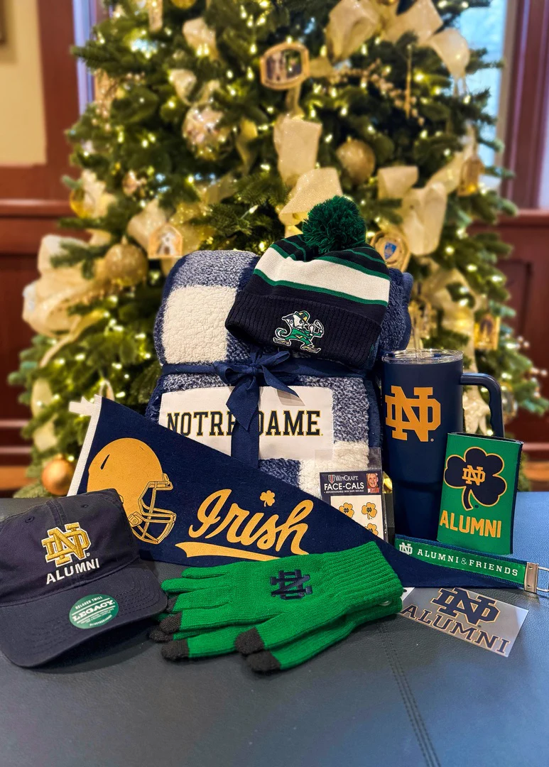 A collection of Notre Dame-themed items is arranged in front of a decorated Christmas tree. The items include a blue and white blanket, beanie, ballcap, gloves,  pennant, reusable cup, drink coozie, keychain, window decal, and face stickers.