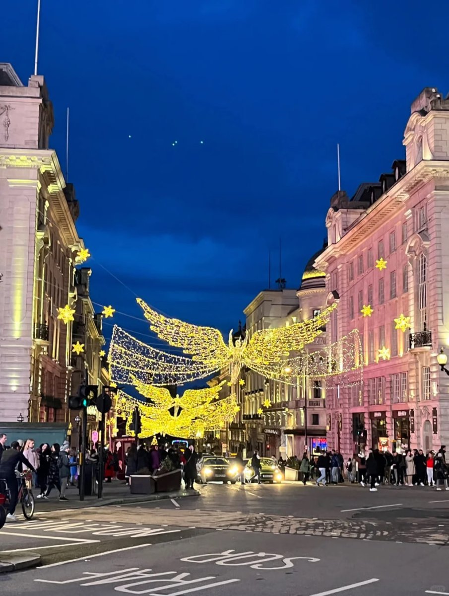 A vibrant city street adorned with dazzling holiday lights shaped like angels, stretching across the bustling roads filled with cars and pedestrians. The backdrop features beautifully lit buildings adding to the festive atmosphere✨