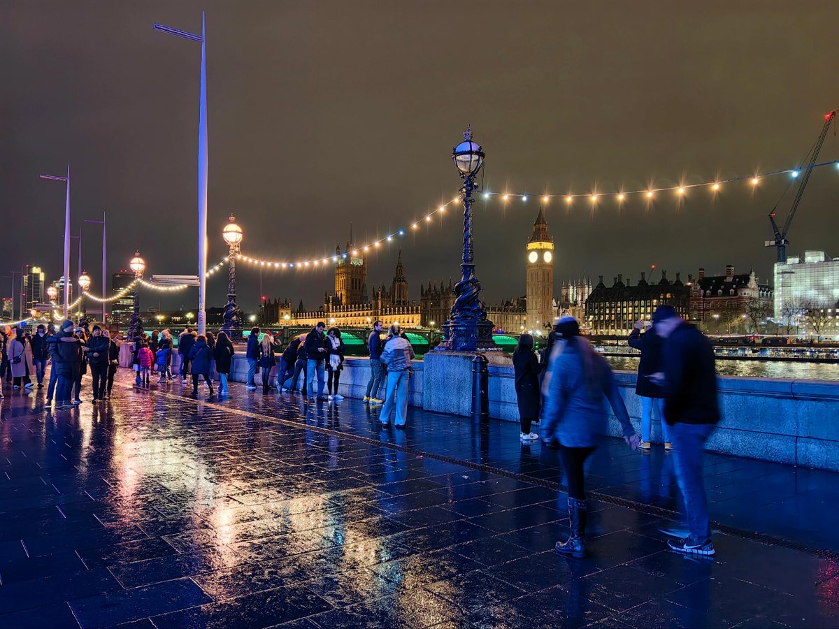 A black cab waits at an intersection near iconic landmarks, with bright city lights reflecting on the wet pavement✨🌟