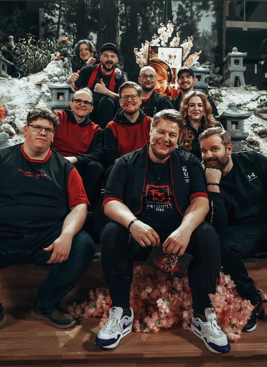 Ein Gruppenbild von den Talents von Friendly Fire, sie sitzen alle zusammen auf einer Treppe, immer 3 Leute pro Stufe und freuen sich alle sehr und Grinsen in die Kamera. Dabei tragen sie alle den Friendly Fire Merch. Der Hintergrund ist japanisch gestaltet. 