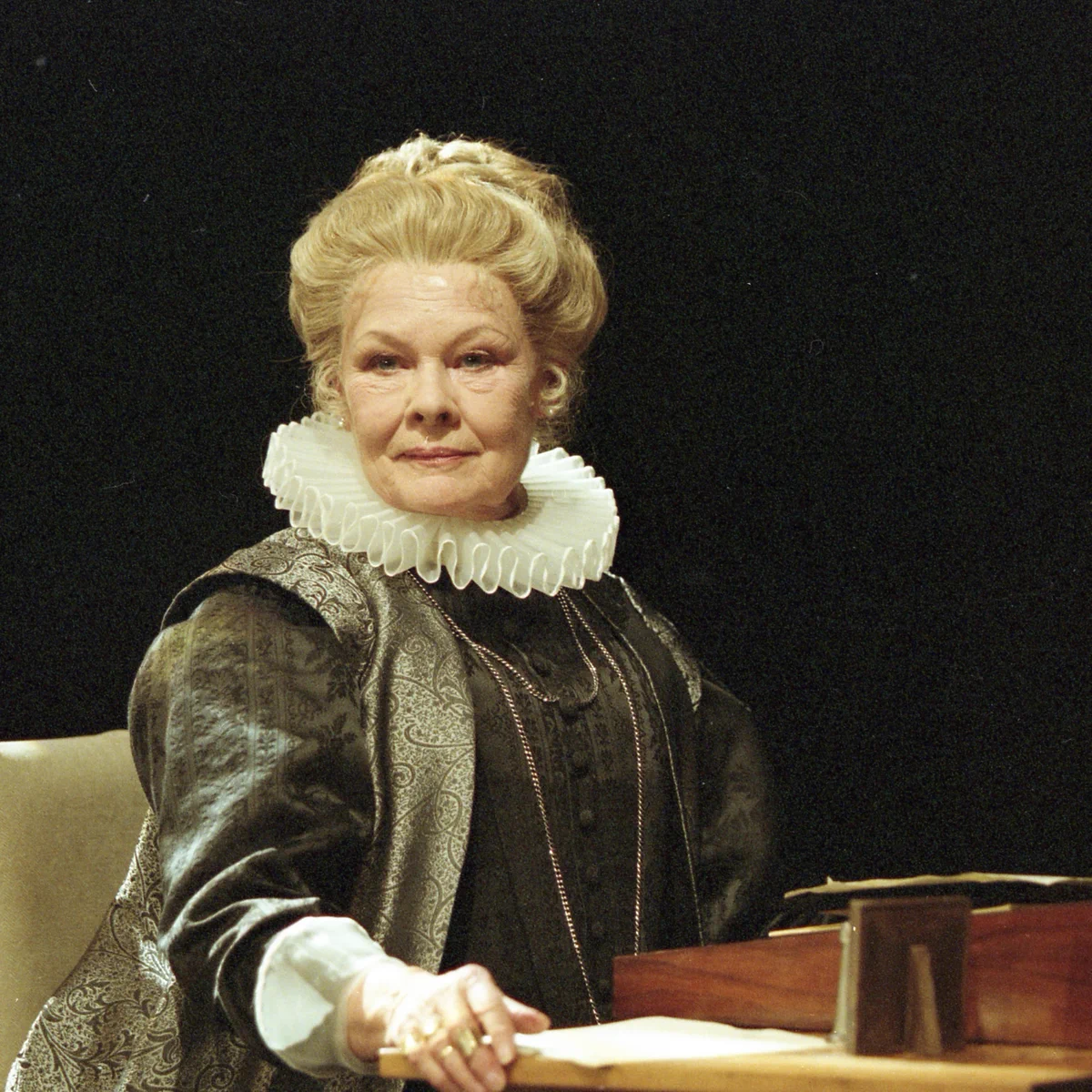 Judi Dench in period costume sits a desk looking forward