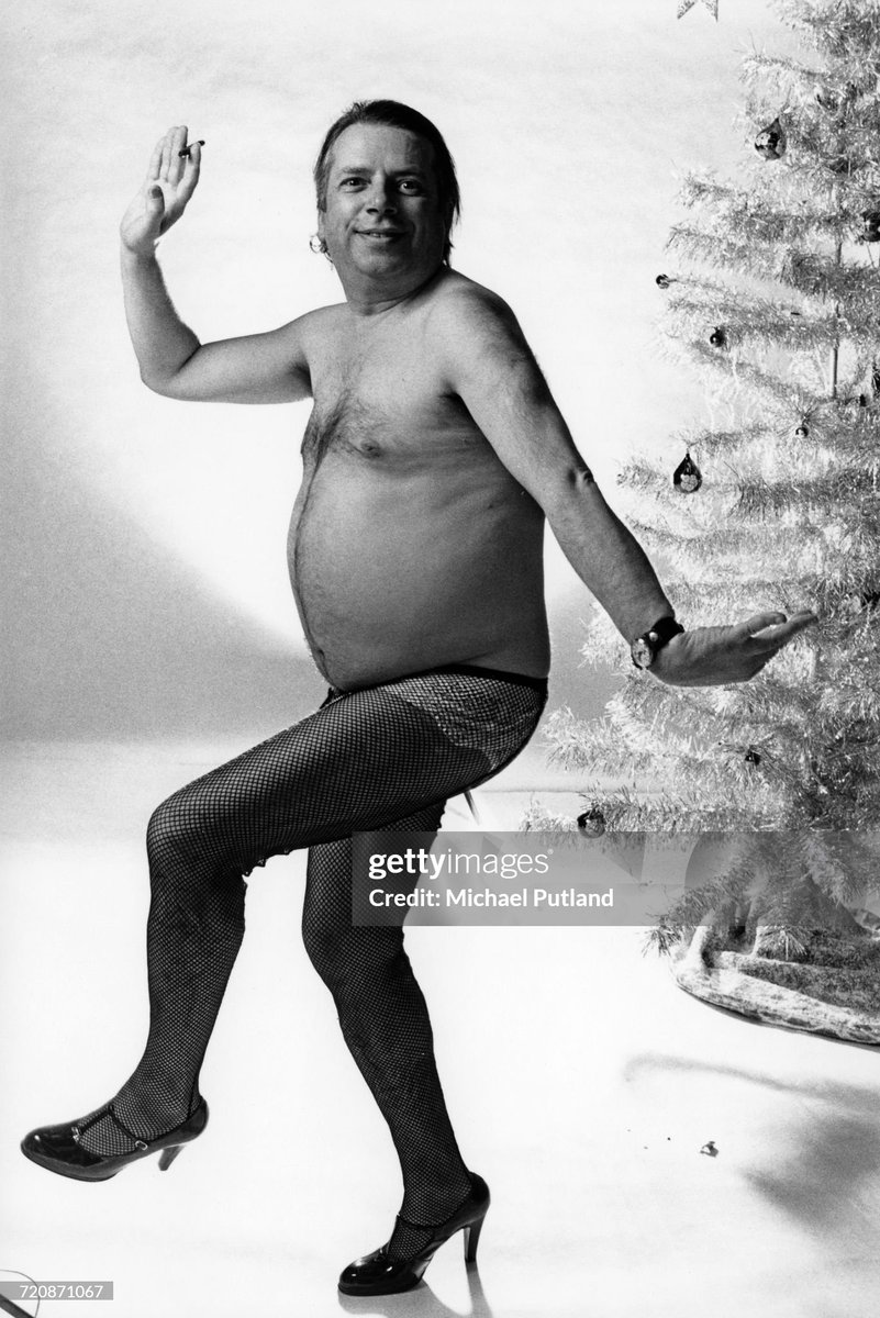 British jazz singer and writer George Melly (1926-2007) pictured bare chested wearing fishnet stockings and high heeled shoes as he poses in front of a synthetic christmas tree (1975)
