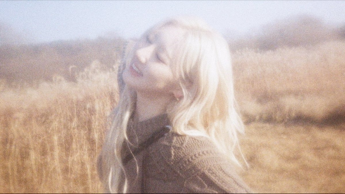 A softly lit, vintage-style photograph of a smiling woman with long blonde hair standing in a sunlit field of tall grass.