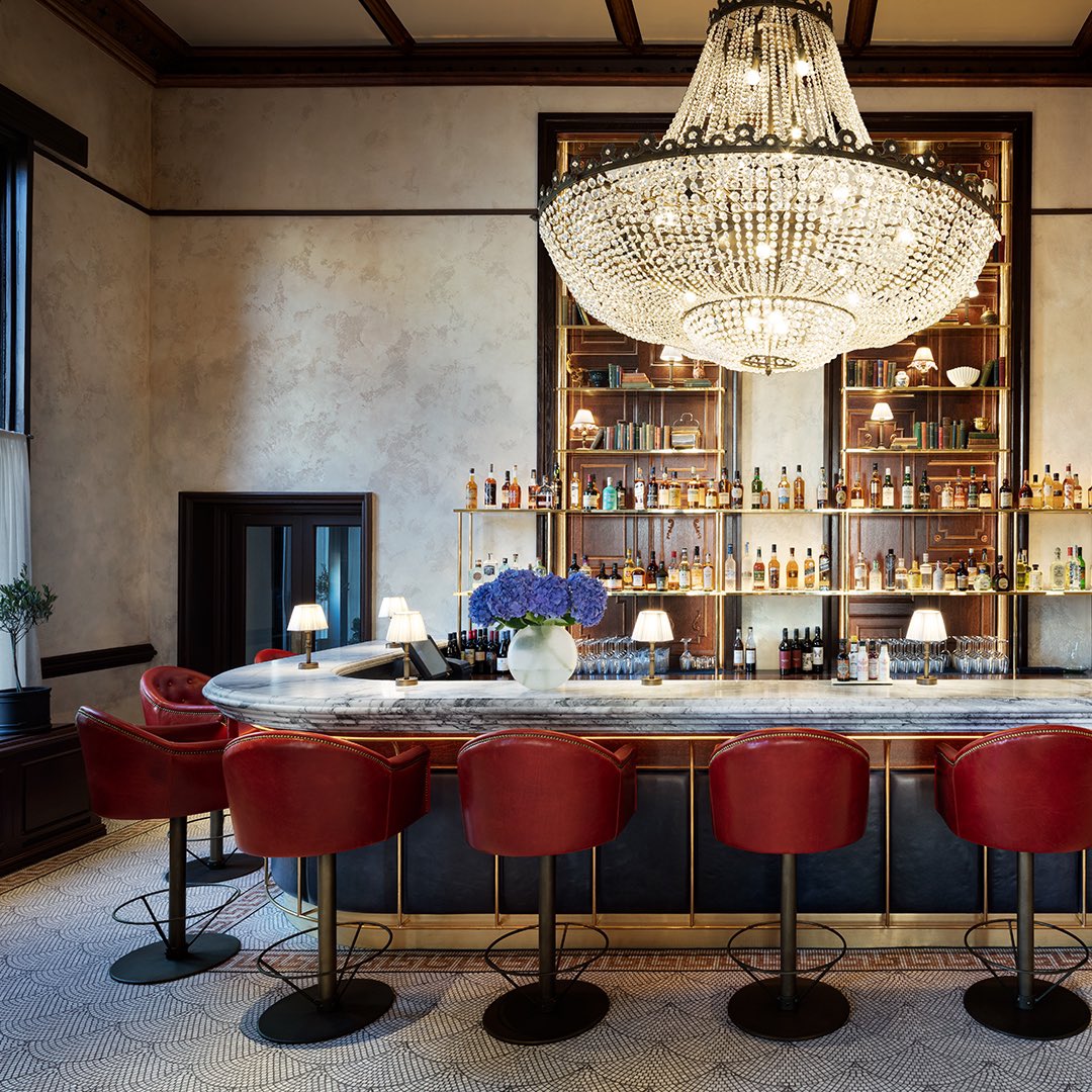 The Alice bar offers an elegant and spacious atmosphere, highlighted by a large chandelier and an open layout. It features a marble bar, leather stools, and a high ceiling, creating a grand ambience.
