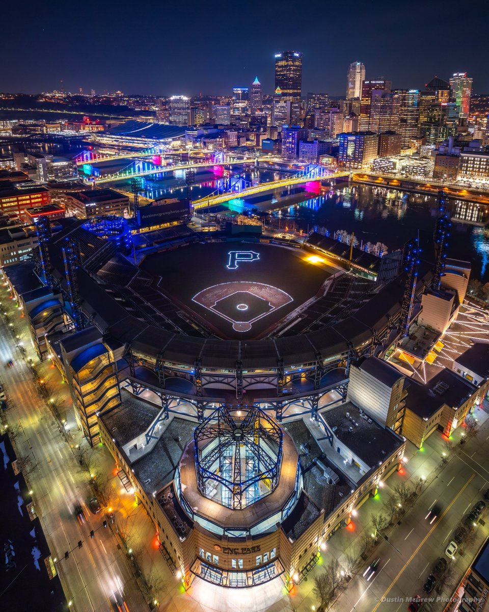 The field inside PNC Park is lit up for the holidays with lights outlining the infield and a Pirates "P" logo in the outfield. Love the colorful sister bridges in the background too! #pittsburgh #mlb #letsgobucs