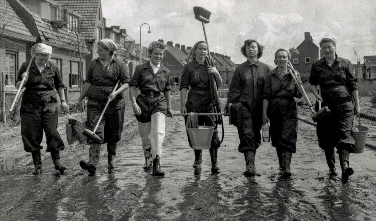 Flood disaster: seven women in overalls and rubber boots, armed with brooms and buckets are on their way to sweep the city clean, Kruiningen, Zuid-Beveland, Zeeland, 1953 - by W.L. Stuifbergen, Dutch.