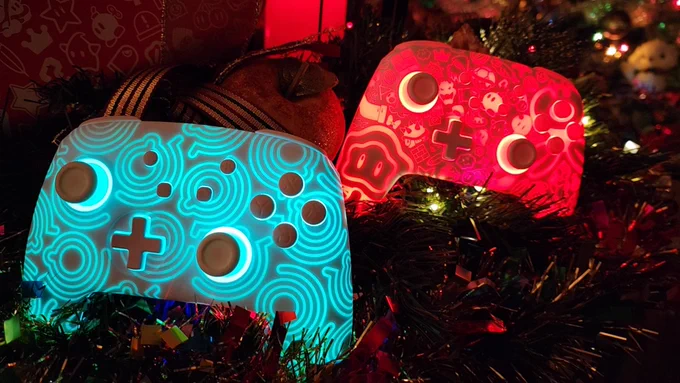 Two wireless Funlab Firefly Pro controllers sitting in a Christmas wreath, in low lighting. The left controller is glowing cyan, with a pattern of runic lines. The right controller is glowing red, with a pattern of Super Mario Wonder characters and items.