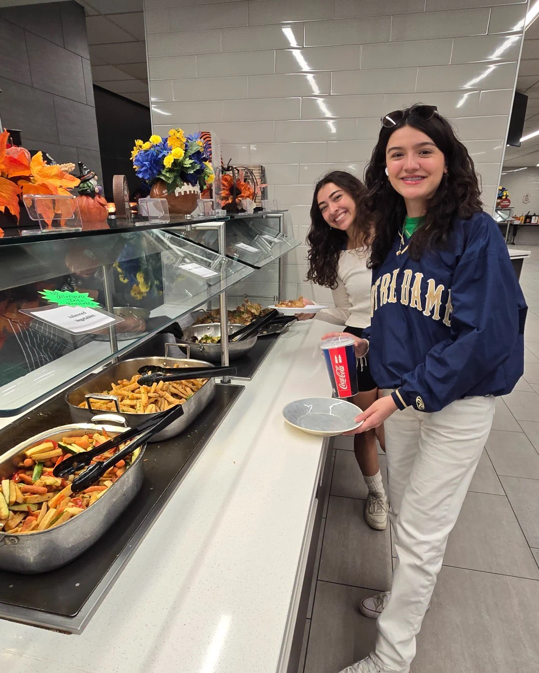 Two students smile while getting food at a buffet line in North Dining Hall. The buffet offers various options, including vegetables and french fries. The student on the right wears a Notre Dame jacket and holds a Coca-Cola cup and plate. Fall decorations, including pumpkins and flowers, adorn the buffet area.