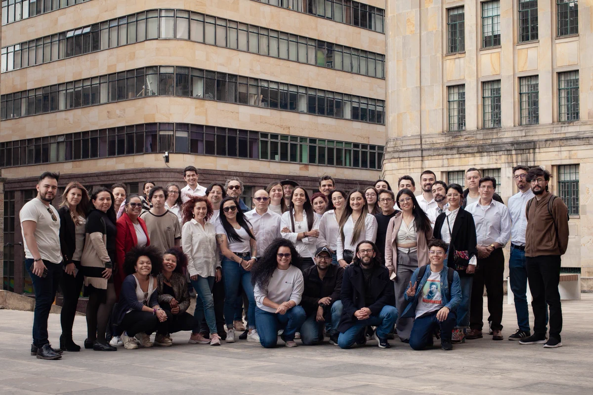 Un grupo diverso de personas posa al aire libre frente a edificios de arquitectura clásica moderna en una ciudad. Todos lucen sonrientes y unidos, reflejando comunidad y colaboración en un entorno profesional. 