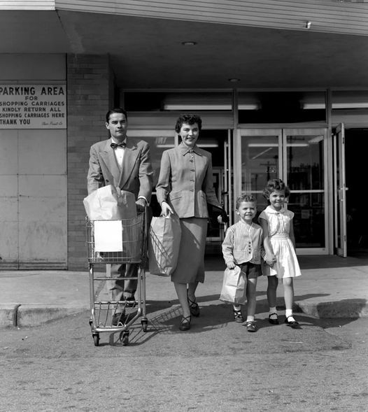 Family walking out of supermarket store pushing grocery cart, 1950s.