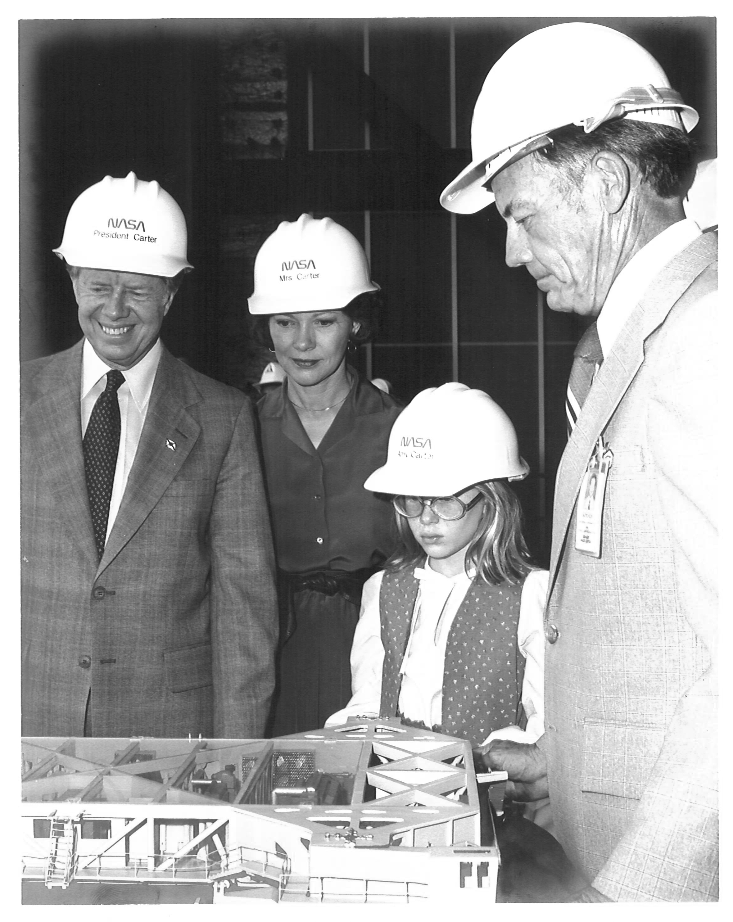 During a tour of Kennedy Space Center, President Jimmy Carter, smiling, with wife Rosalynn and daughter Amy, listen to center director Lee R. Scherer discuss a crawler-transporter model on the table in front of them. They are wearing hard hats with the NASA "worm" logo, and each Carter family member's name. Credit: NASA 