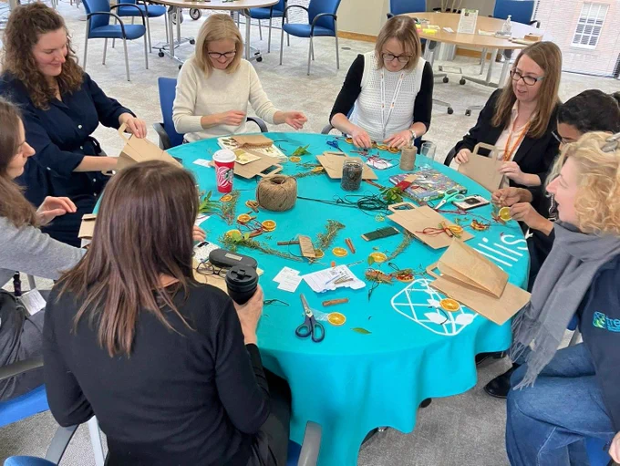 8 people sit around a round table with a turquoise table cloth which has crafting supplies scattered over it. 
