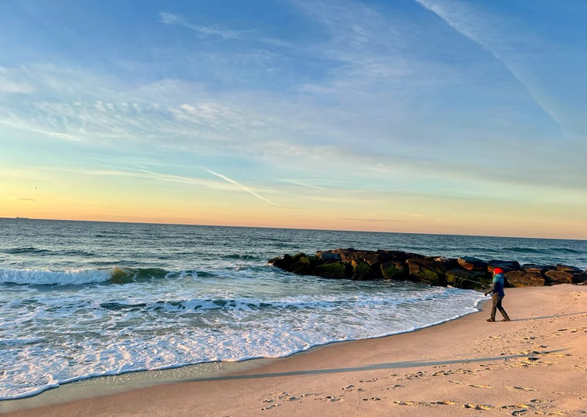 On the beach where the waves are gently lapping, facing the rising sun, the shadow of the fisherman is lengthened by the sun, outlining the tranquil beauty of the early morning.