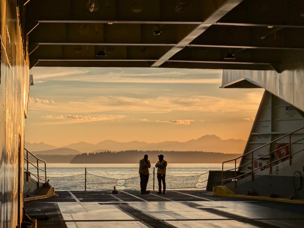 Two people standing at the end of the car deck of a ferry, silhouetted by the sunrise with calm water and mountains behind them.