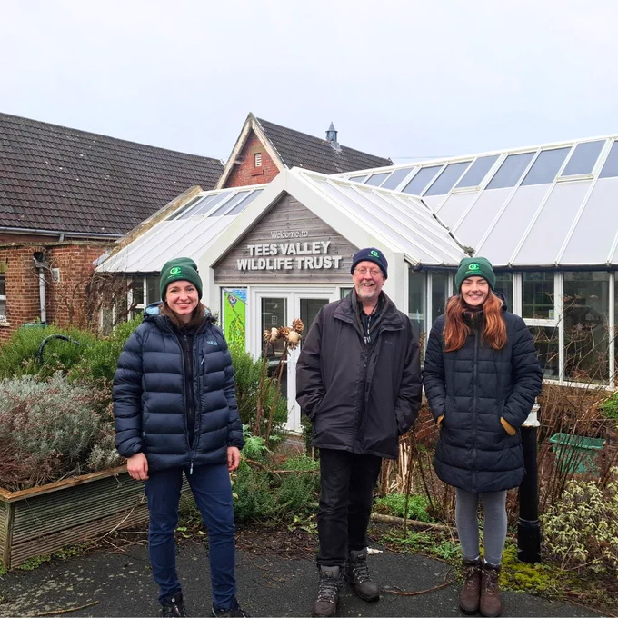 Three people stand outside a building with a sign saying Tees Valley Wildlife Trust. They are wearing hats with the words Open Country and are smiling.