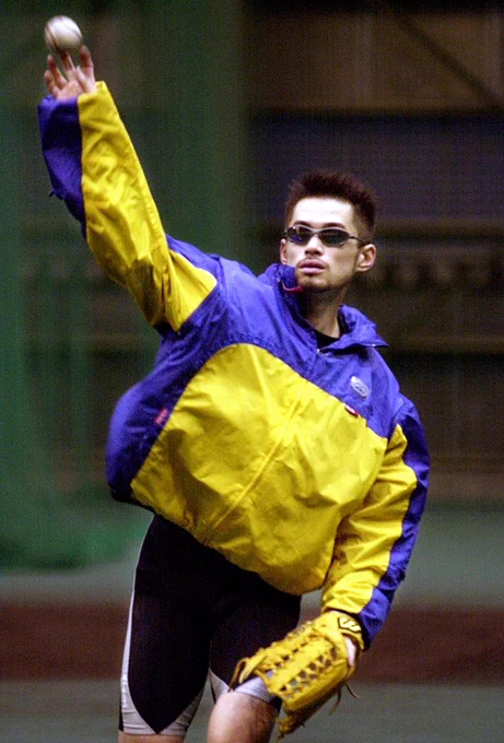 Photo of Ichiro Suzuki throwing a baseball while wearing a yellow and purple windbreaker and bike shorts, looking very cool. 