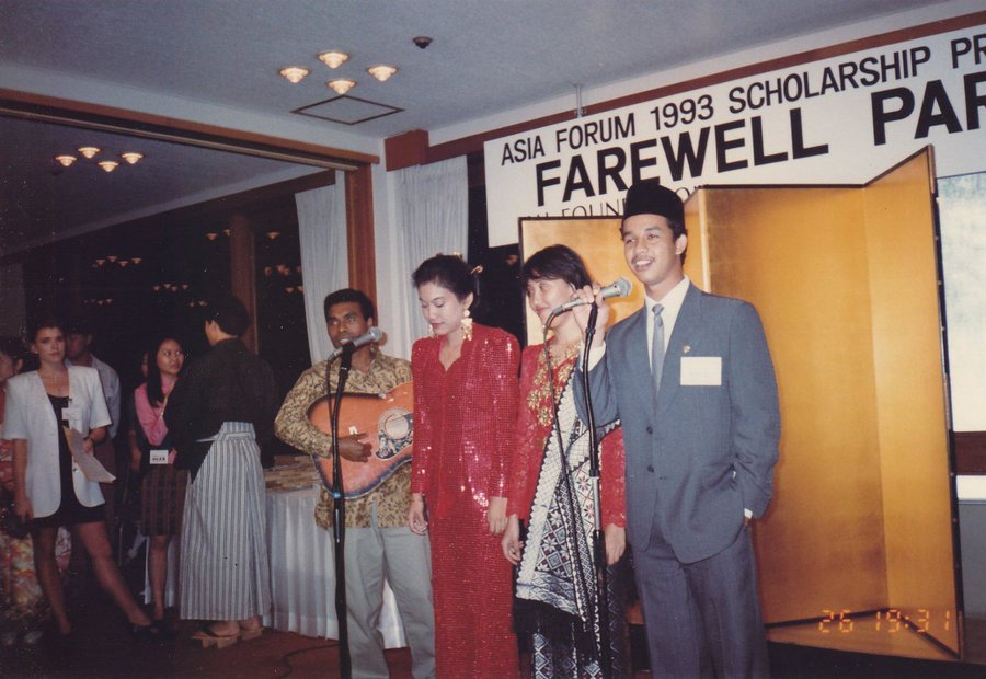 Foto Anies Baswedan muda bersetelan jas abu-abu dan mengenakan peci sedang bernyanyi memegang mikrofon dalam acara perpisahan Asia Forum 1993 Scholarship Program. Di sampingnya, seorang pria memainkan gitar, serta dua perempuan berpakaian tradisional berdiri di tengah. Latar belakang menunjukkan dekorasi dengan tulisan ‘Farewell Party’.