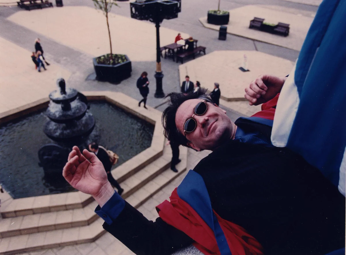 Tony Slattery, wearing sunglasses and the Rector's robes from the University of Dundee's Students' Association, DUSA, leans over the edge of a balcony above City Square, Dundee