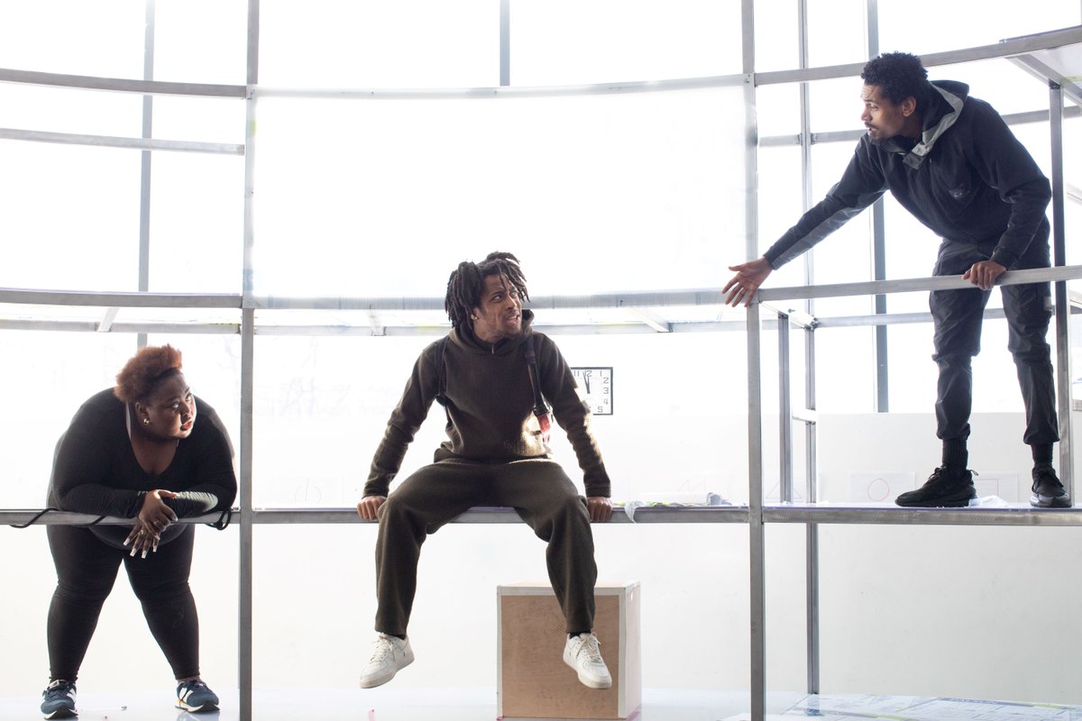 Pig Heart Boy in rehearsals. Photo by Ali Wright. Three people are interacting on a scaffold structure. One person leans over a bar lower down, one in the centre sits on a platform and the highest one stands on a platform and leans down as they speak to the others.