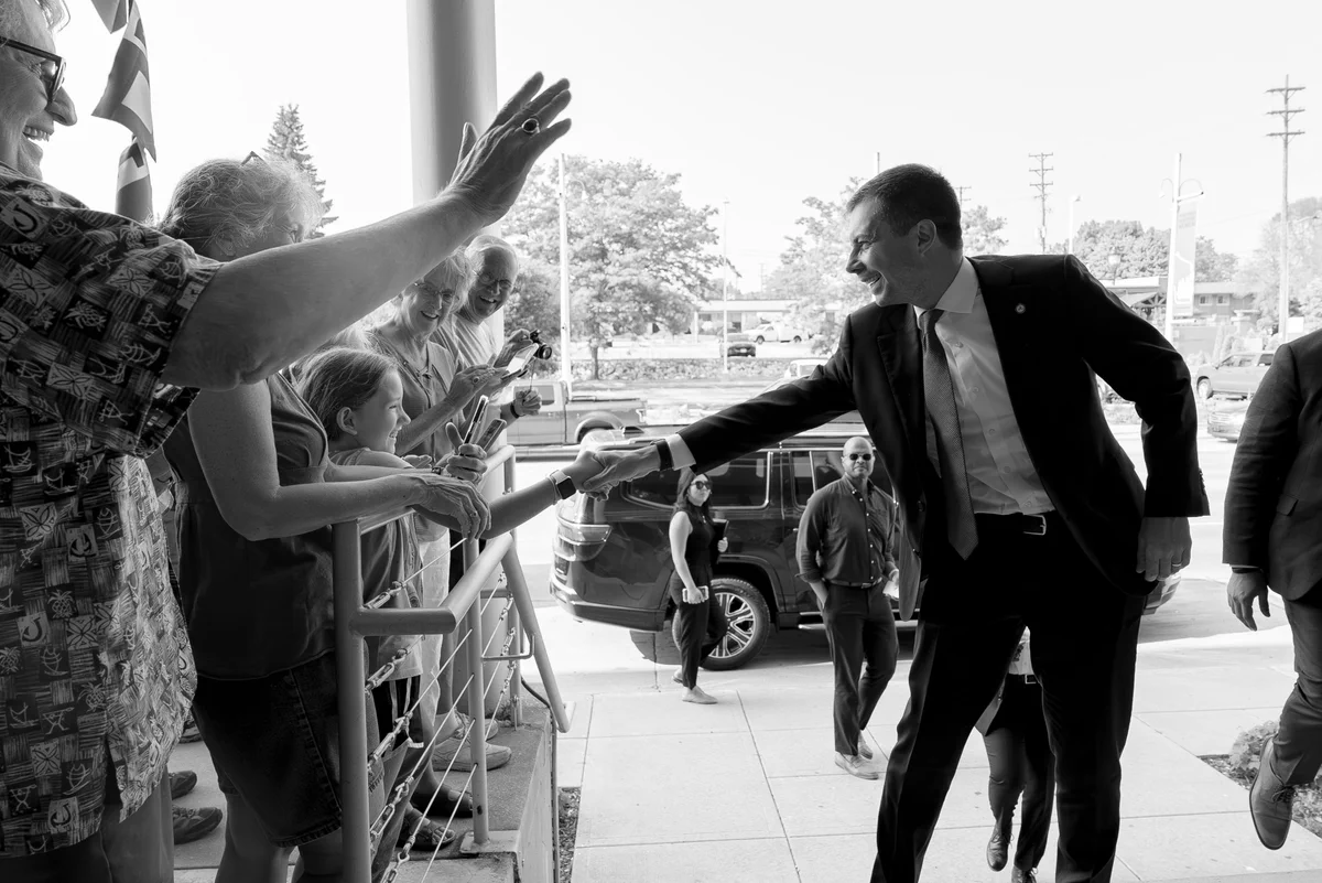 Black-and-white image of Secretary Pete Buttigieg shaking hands with a child at an event. 