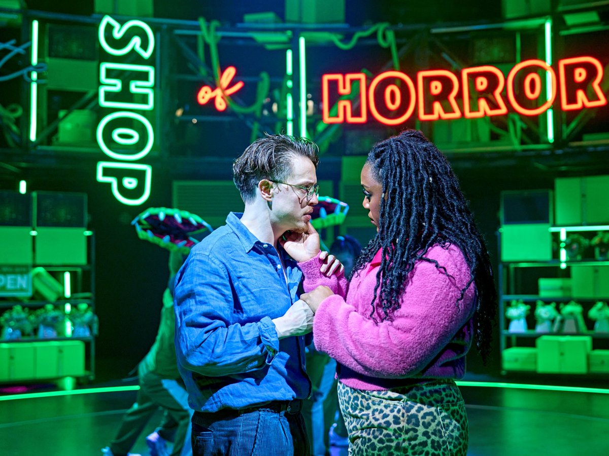 Colin Ryan (Seymour), Georgina Onuorah (Audrey) and members of the company in Little Shop of Horrors. Photo Manuel Harlan. Two people stand close together and look ugently at one another. One holds the other's face in their hands. Behind in neon reads SHOP OF HORROR