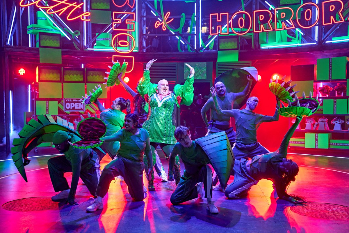Sam Buttery (The Plant (Audrey II)) and members of the company in Little Shop of Horrors. Photo Manuel Harlan. A person wearing green sequins holds their arms in the air and others surrounding them sprawl out, dressed as leaves and plant buds.
